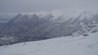 4K aerial stock footage flying over snowy summit toward a wooded valley, Chugach Mountains, Alaska Aerial Stock Footage | AK0001_1171