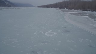 AK0001_1272 - 4K aerial stock footage following an icy river and tree lined, snowy riverbank, Butte, Alaska