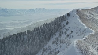 AX125_197 - 5.5K aerial stock footage orbit snowdrifts blowing from a mountain peak with evergreens in winter, Oquirrh Mountains, Utah