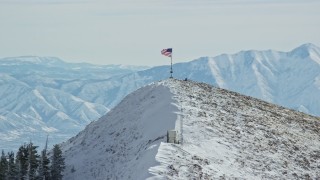 AX125_210E - 5.5K aerial stock footage approach American flag waving atop a snowy peak in winter, Oquirrh Mountains, Utah